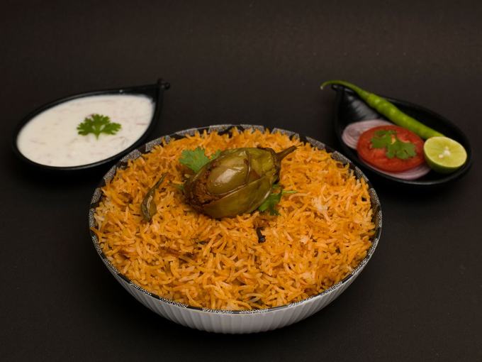 Biryani in a steel dish with raita, lime, tomato and green chilli alongside.
