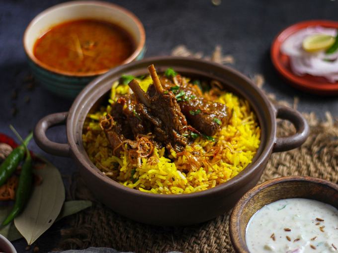 Lamb biryani on saffron rice in a dark bowl, with raita and curry alongside.