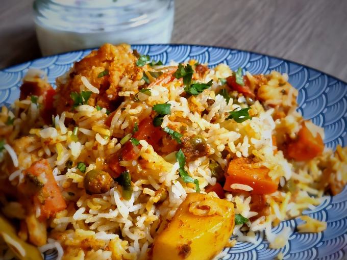 A colourful vegetable biryani on a patterned plate with a pot of yoghurt.