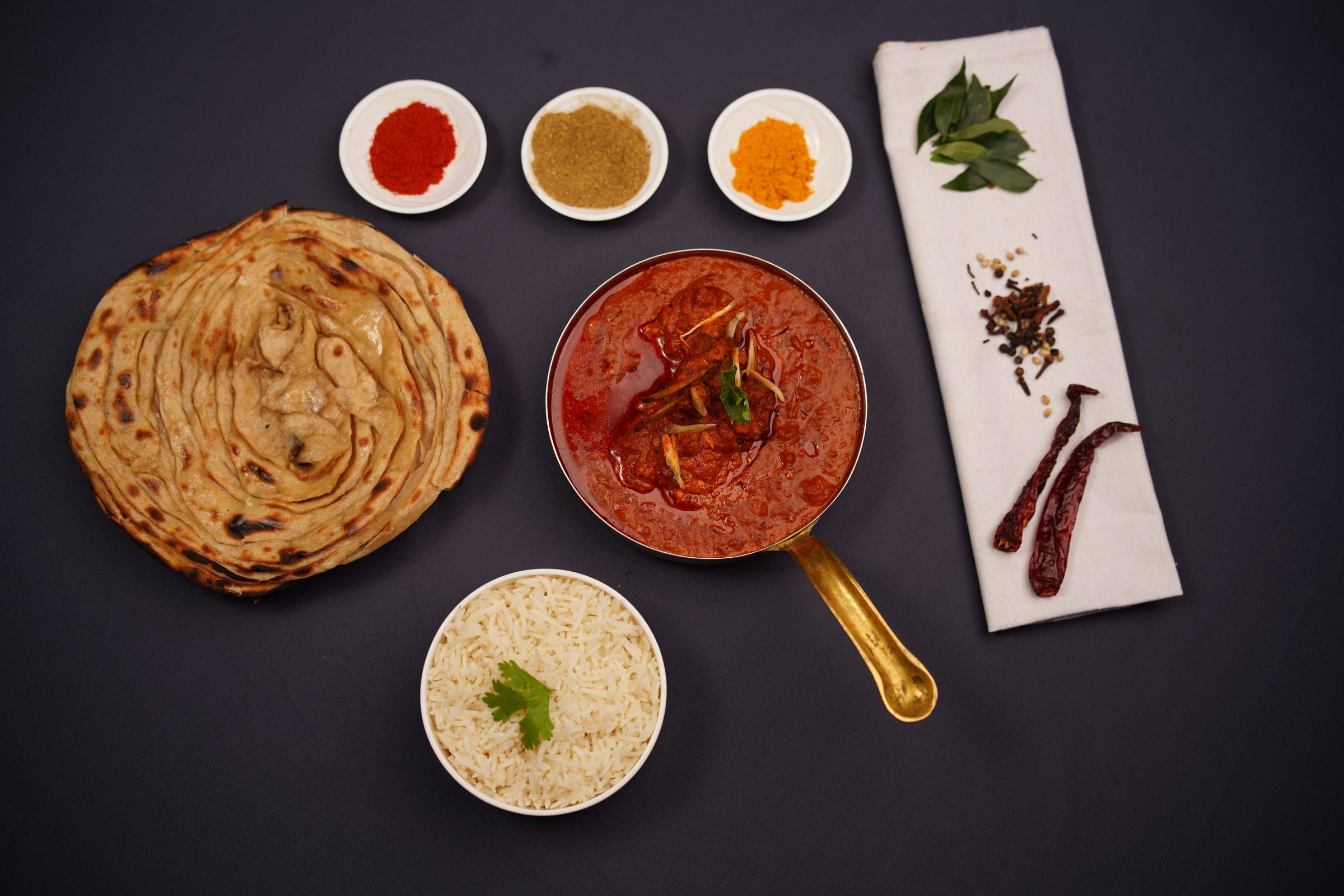 An overhead spread on dark slate: a copper karahi of rich curry, flaky paratha, a bowl of rice and a row of whole spices.