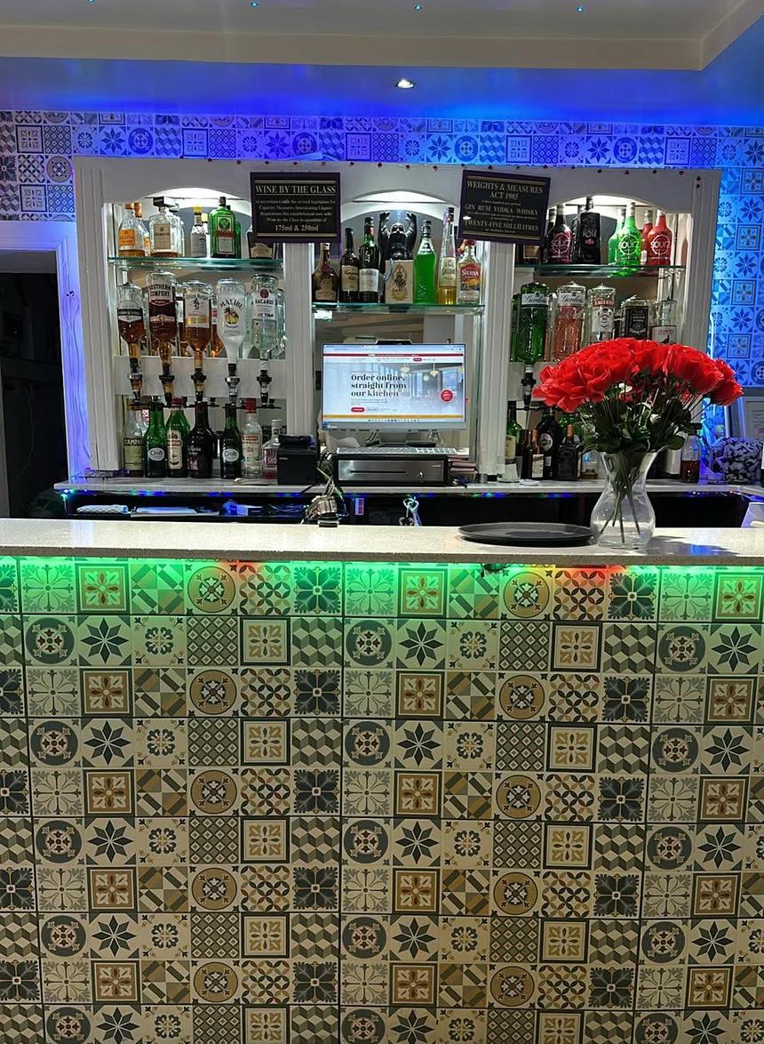 The bar: a counter of patterned tiles lit green from beneath, shelves of spirits against blue-lit tiles, and a vase of red roses.
